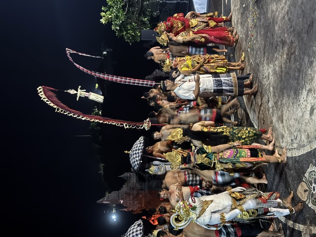 A Balinese community gathered at night in ceremonial dress, with performers, temple structures, umbrellas and penjor rising above the crowd.