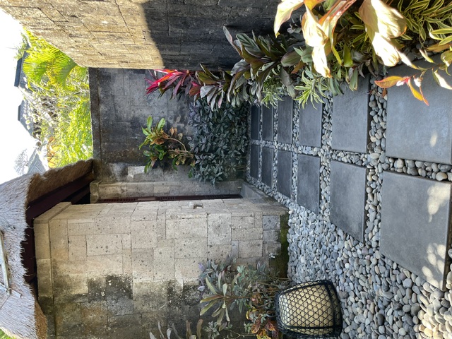 A quiet stone path through a Balinese courtyard, bordered by white pebbles, tropical plants and a traditional thatched structure.